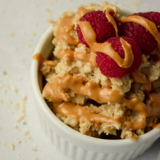 Small white ramekin of peanut butter oat bran with a peanut butter drizzle and raspberries